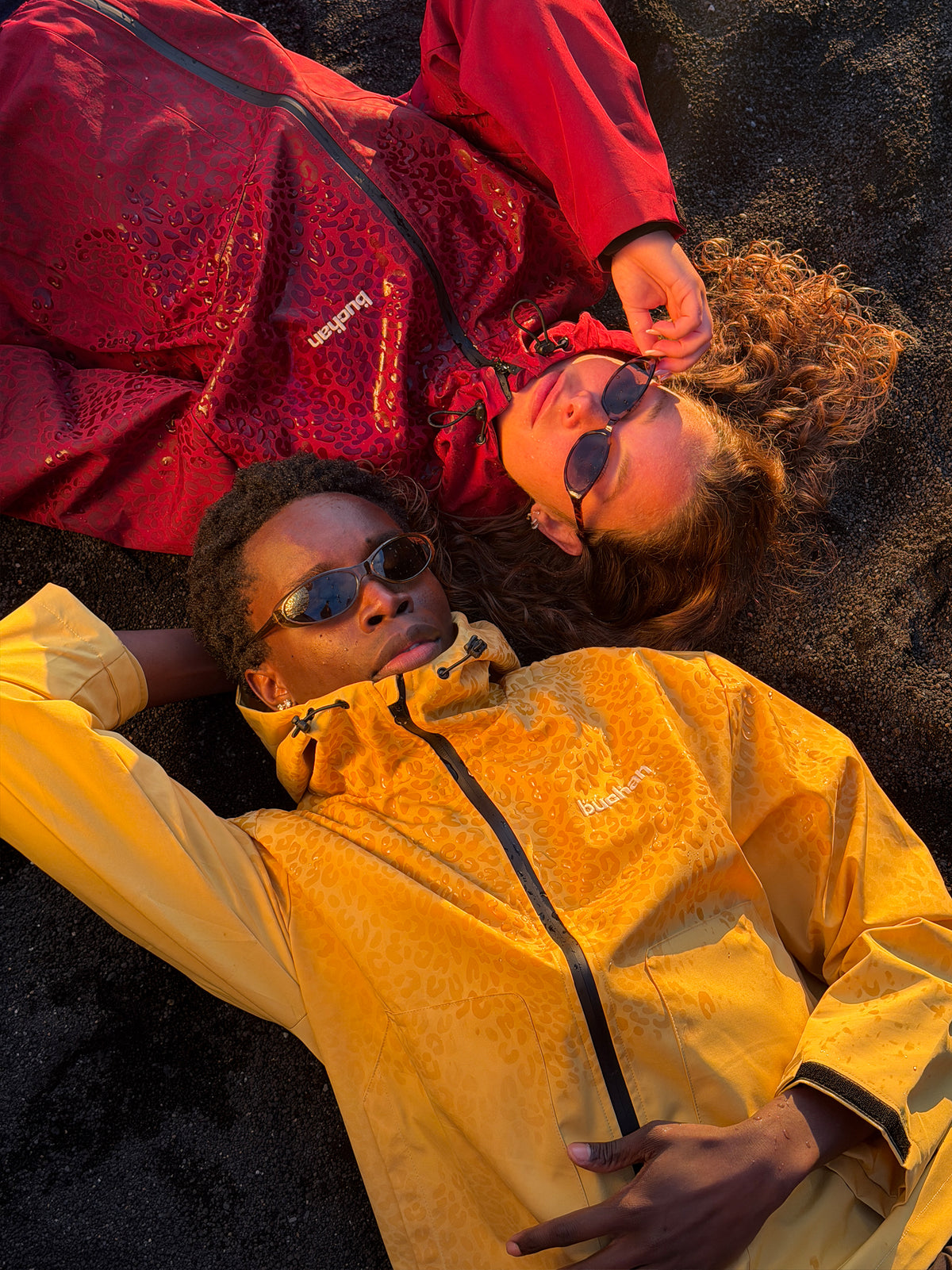 Two models lying on black sand, one in the water reactive cheetah rain jacket in yellow, print visible