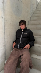 Model sitting on concrete stairs in the reversible Buchan fleece in khaki, worn on the black nylon side
