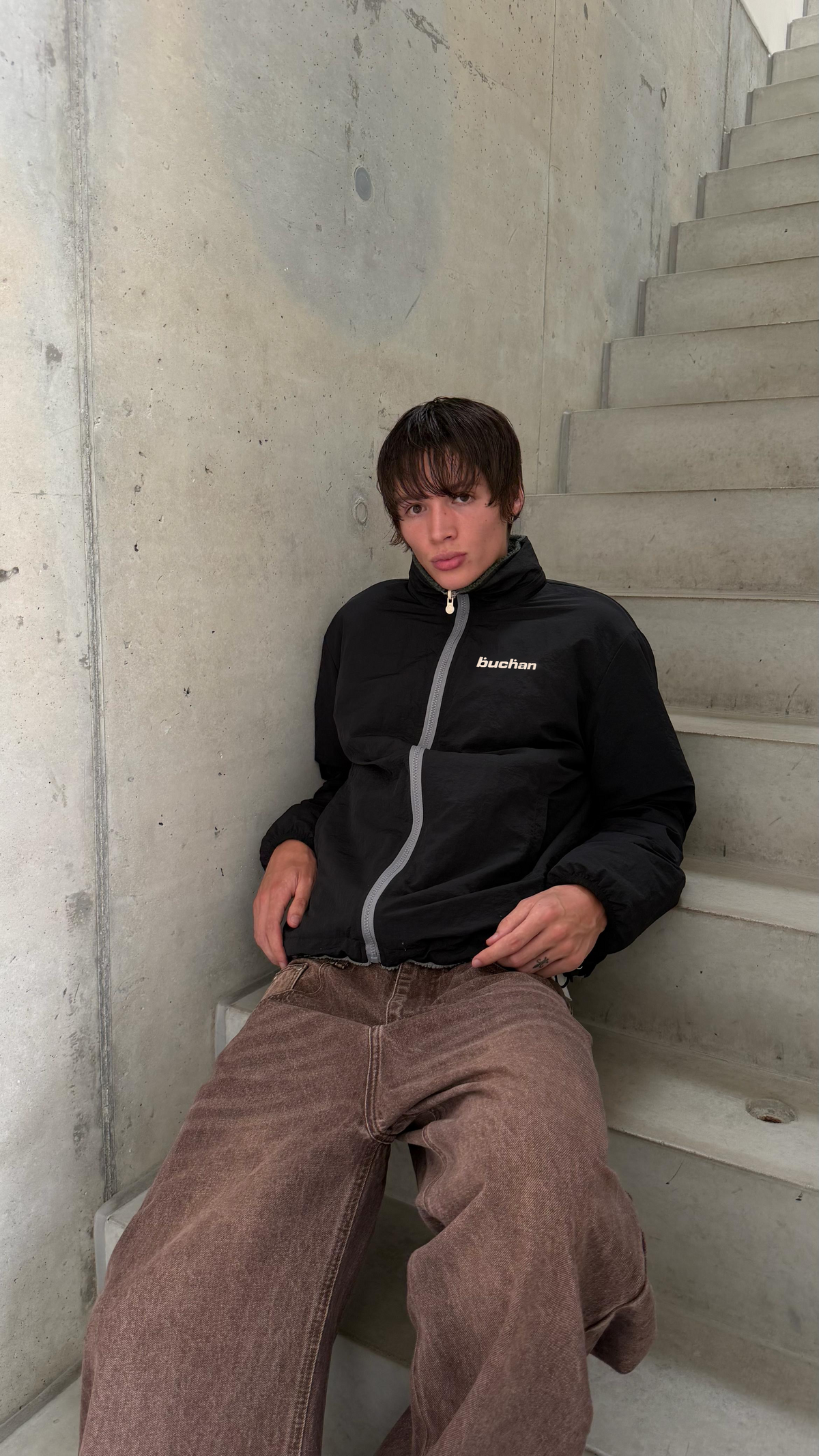 Model sitting on concrete stairs in the reversible Buchan fleece in khaki, worn on the black nylon side