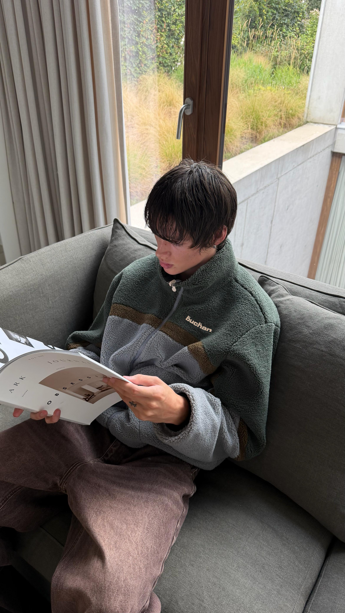 Model reading on a sofa in the reversible Buchan fleece in khaki, green and grey sherpa side out