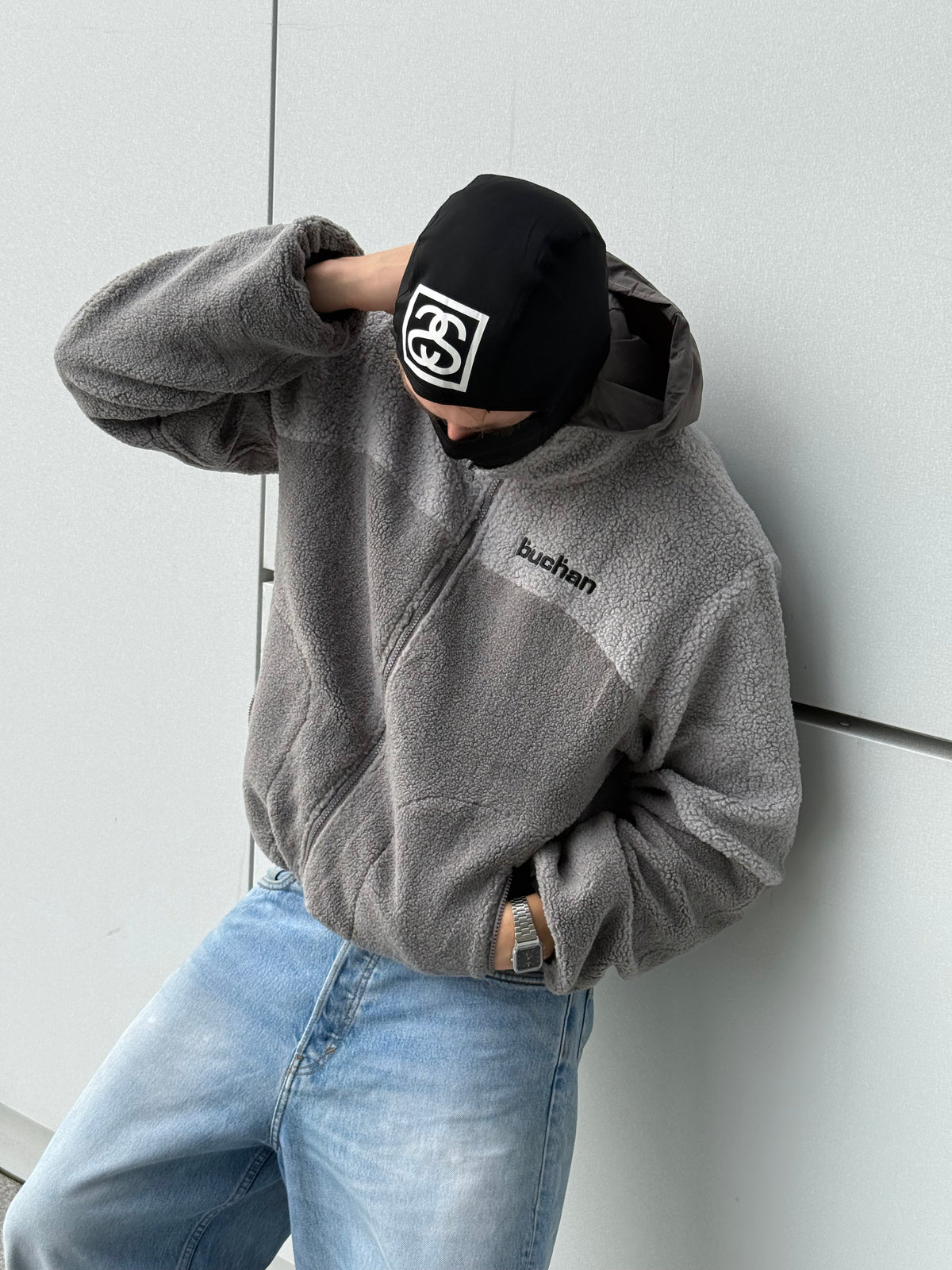 Model leaning against a wall in the reversible Buchan fleece in grey, sherpa side with the hood up