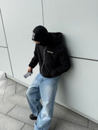 Model leaning on a wall in the reversible Buchan fleece in black worn on the black nylon side, hood up