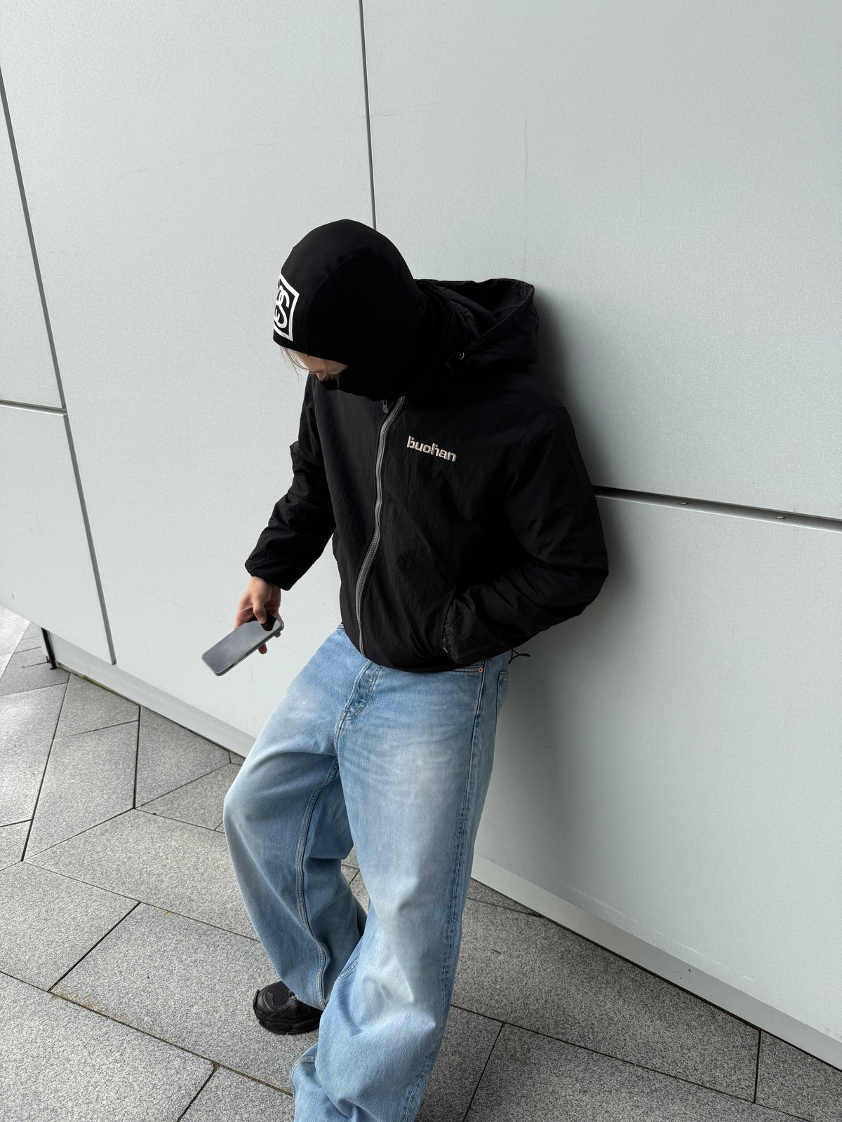 Model leaning on a wall in the reversible Buchan fleece in black worn on the black nylon side, hood up