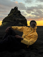 Model sitting on black sand at sunset in the water reactive cheetah rain jacket in yellow