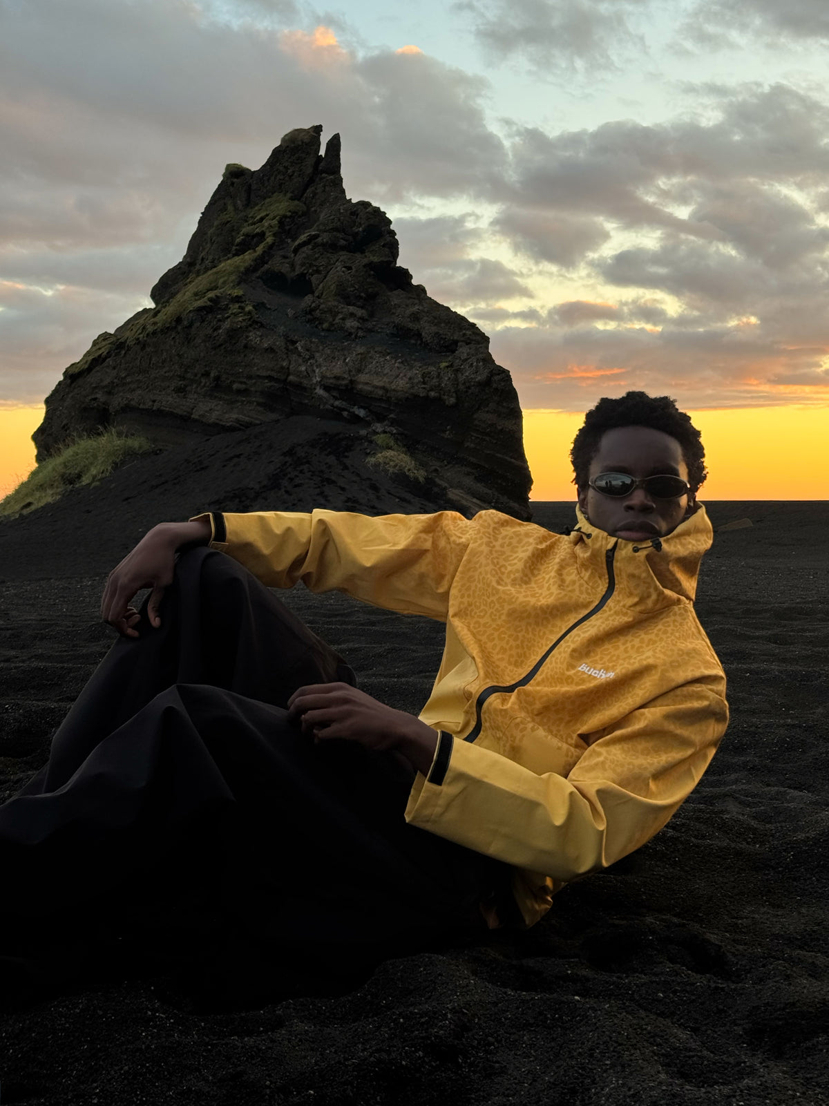 Model sitting on black sand at sunset in the water reactive cheetah rain jacket in yellow